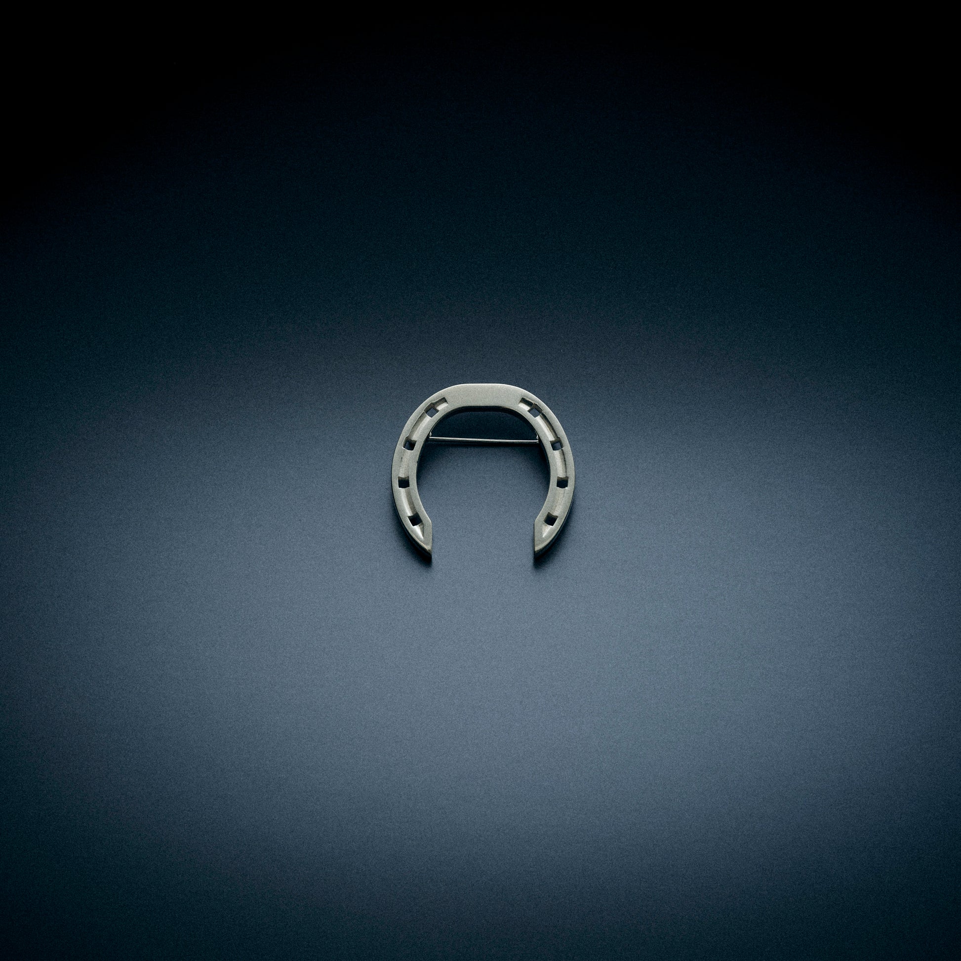 A sleek horseshoe-shaped brooch without stones, displayed on a dark, textured background. The brooch features a minimalist design with a horizontal clasp on the back.