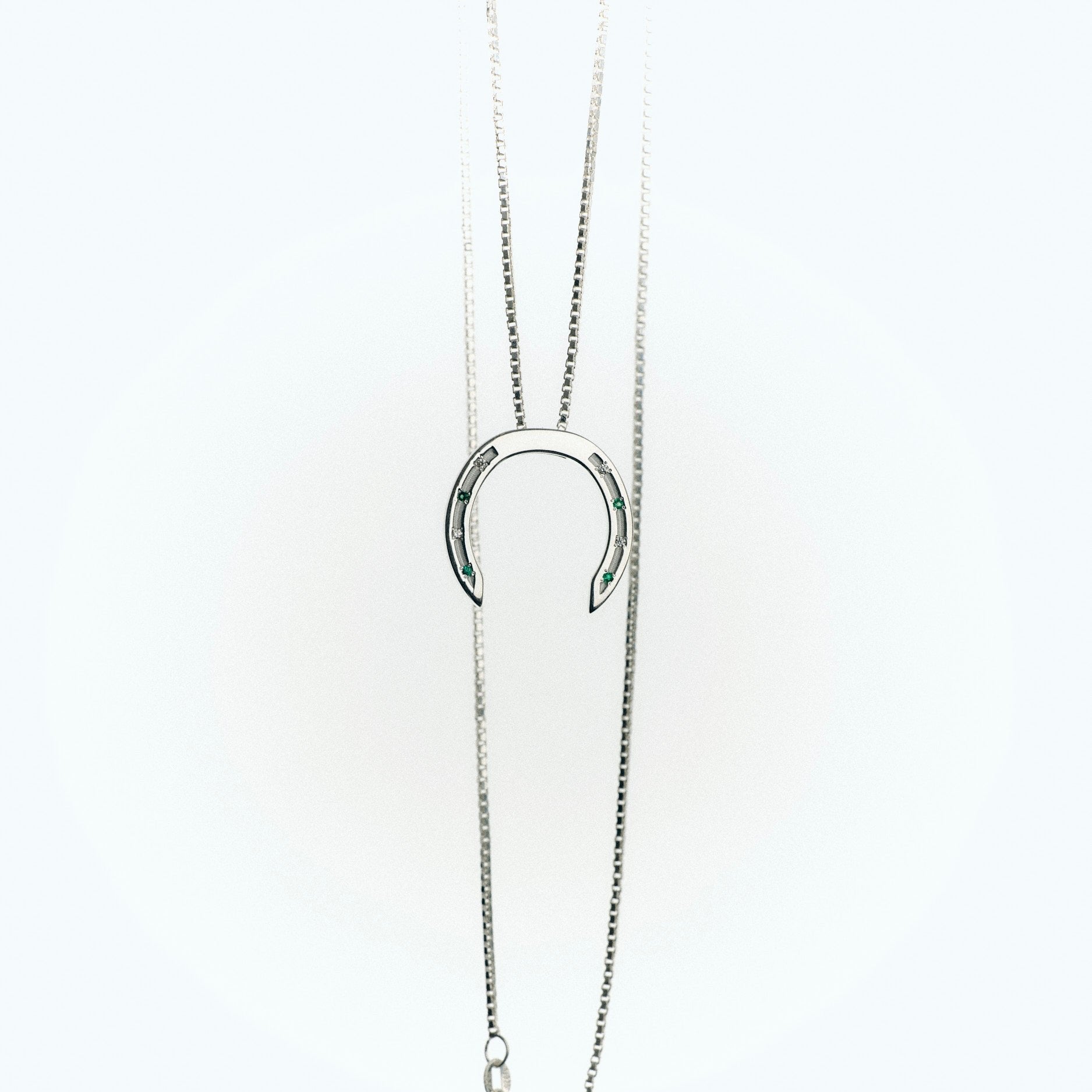 A delicate silver necklace with a horseshoe pendant, featuring small green emeralds and white diamonds embedded in the design. The chain is elegantly draped, with a clasp visible at the bottom, set against a light background.