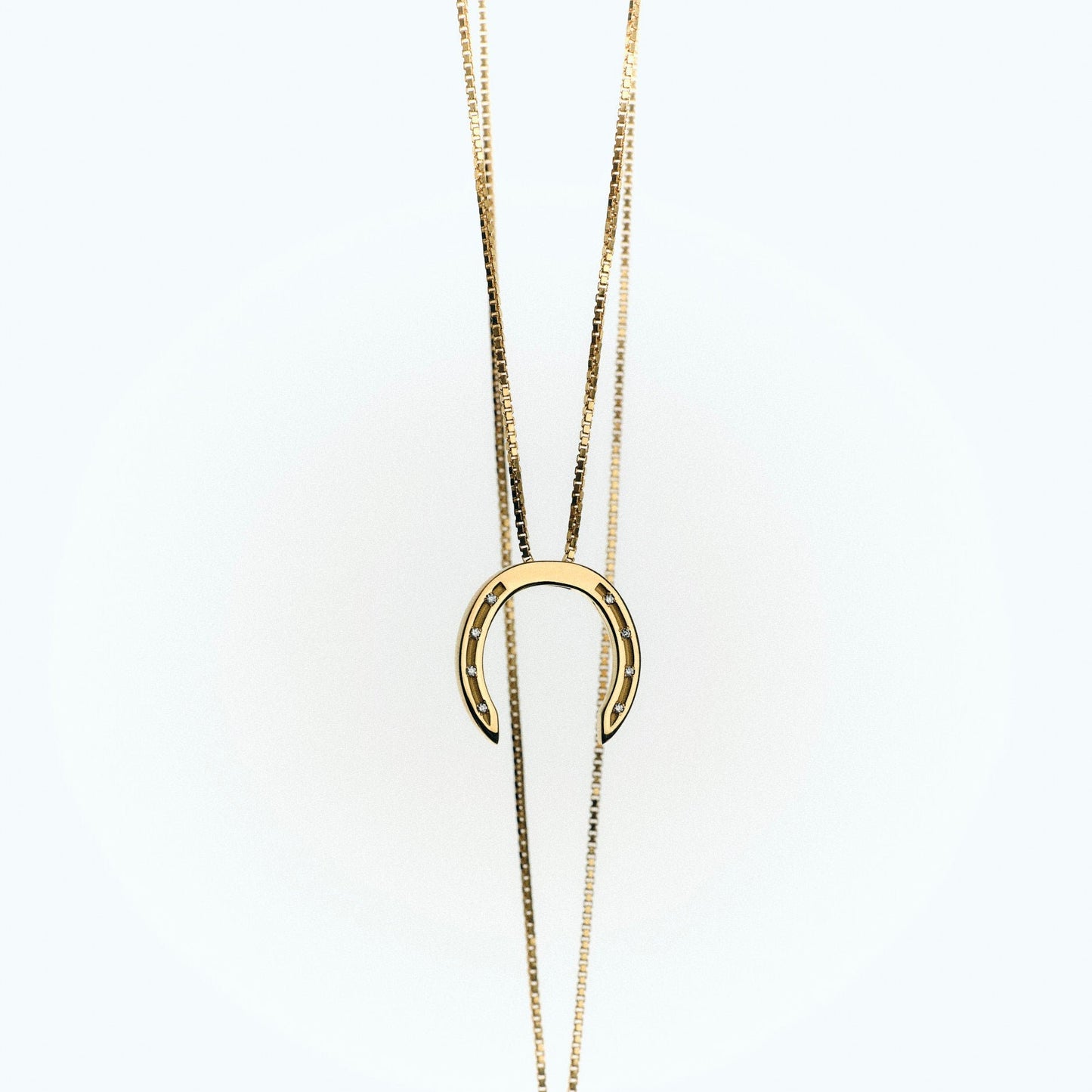 A delicate gold necklace with a horseshoe pendant, featuring small diamonds embedded in the holes, set against a light background.