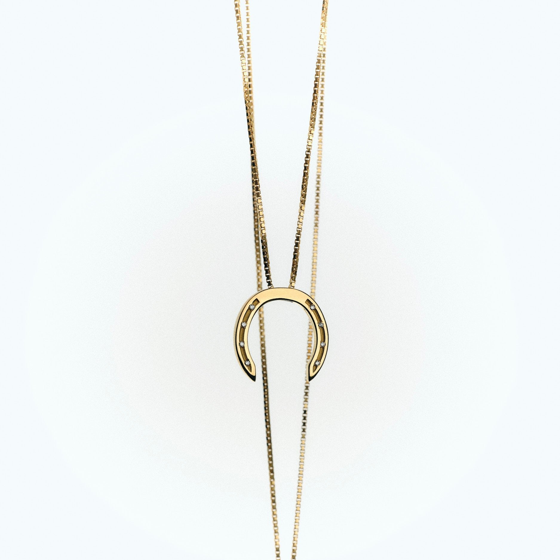 A delicate gold necklace with a horseshoe pendant, featuring small diamonds embedded in the holes, set against a light background.